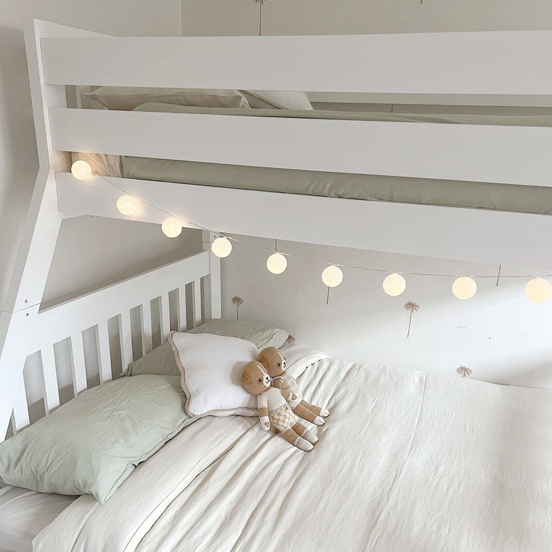 Kourtney Hardwood Triple Bunk Bed with Drawers in White featuring glowing lantern lights and knitted teddy bears in a minimalist nursery room with palm-tree motif wallpaper.