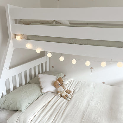 Kourtney Hardwood Triple Bunk Bed with Drawers in White featuring glowing lantern lights and knitted teddy bears in a minimalist nursery room with palm-tree motif wallpaper.