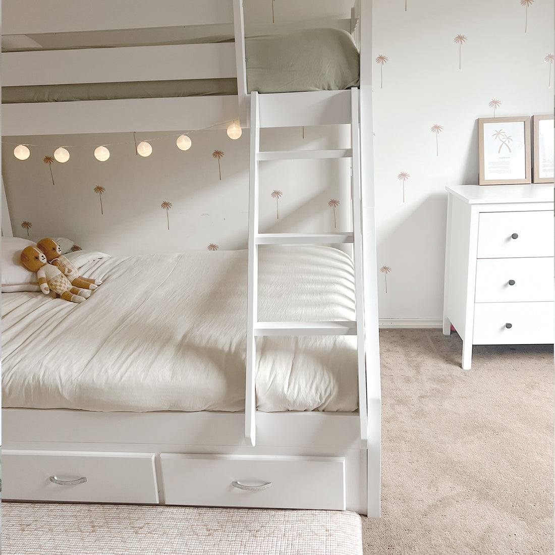 Kourtney Hardwood Triple Bunk Bed with Drawers in White, featuring built-in storage, globe lights beneath upper bunk, set in a cozy children's room with palm motif walls and matching décor.