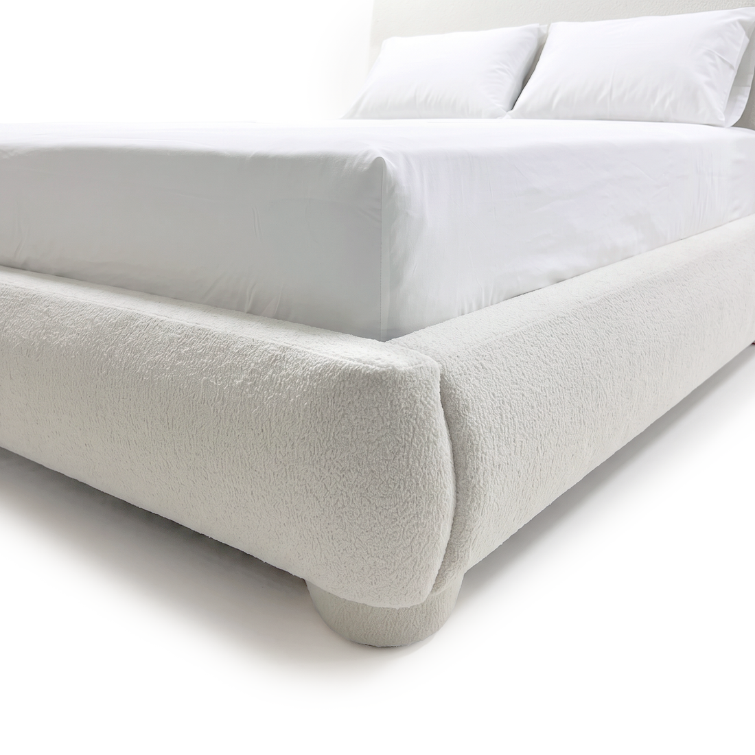 A close-up view of the white bouclé bed frame showing the rounded corner detail, upholstered low footboard, and a matching rounded foot on a white background.