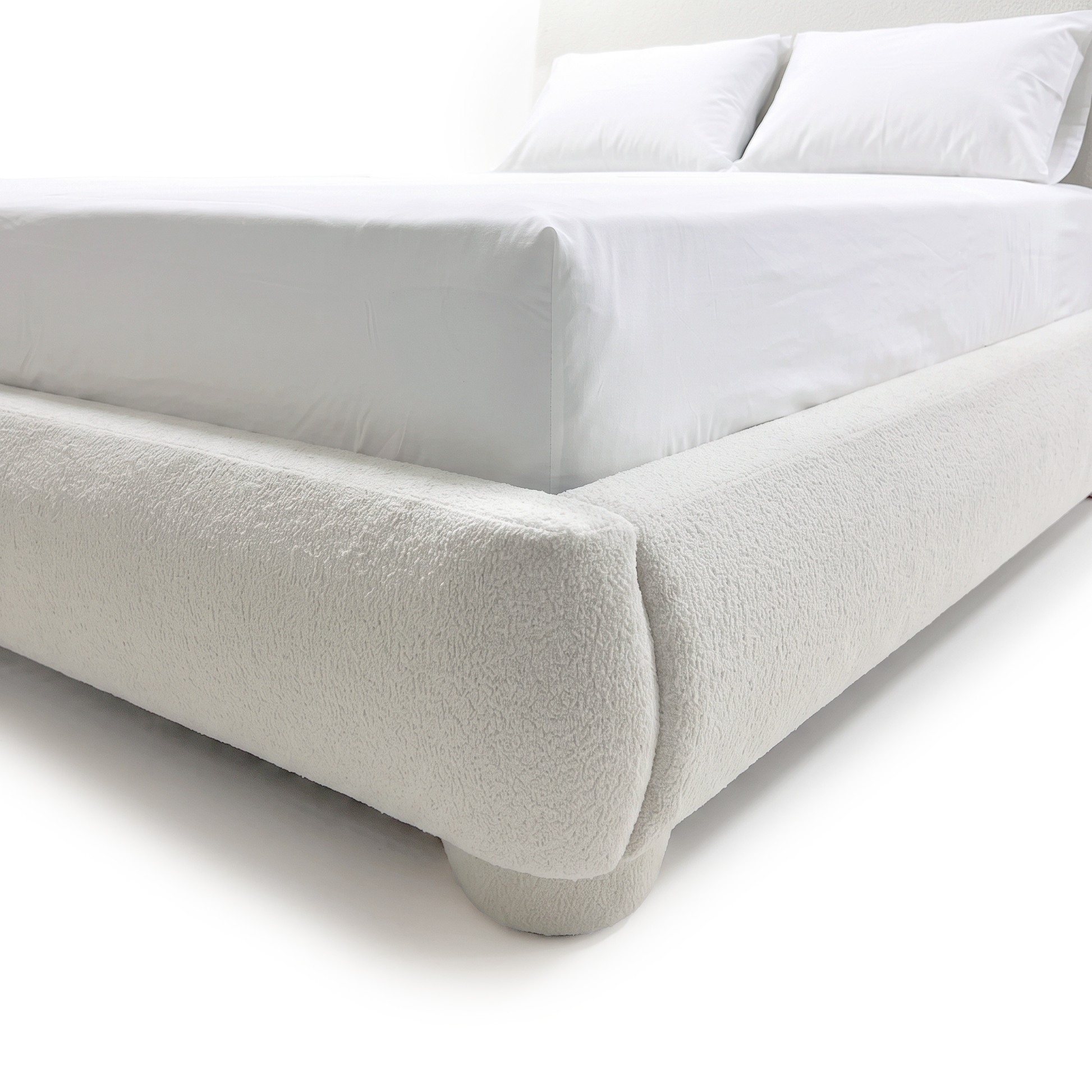 A close-up view of the white bouclé bed frame showing the rounded corner detail, upholstered low footboard, and a matching rounded foot on a white background.