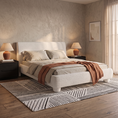 A modern bedroom featuring the Premium Cream Boucle Bed with oversized upholstered legs and a deep-padded headboard, styled with beige and rust-toned bedding, flanked by dark nightstands with lamps on a geometric patterned rug over hardwood floors.