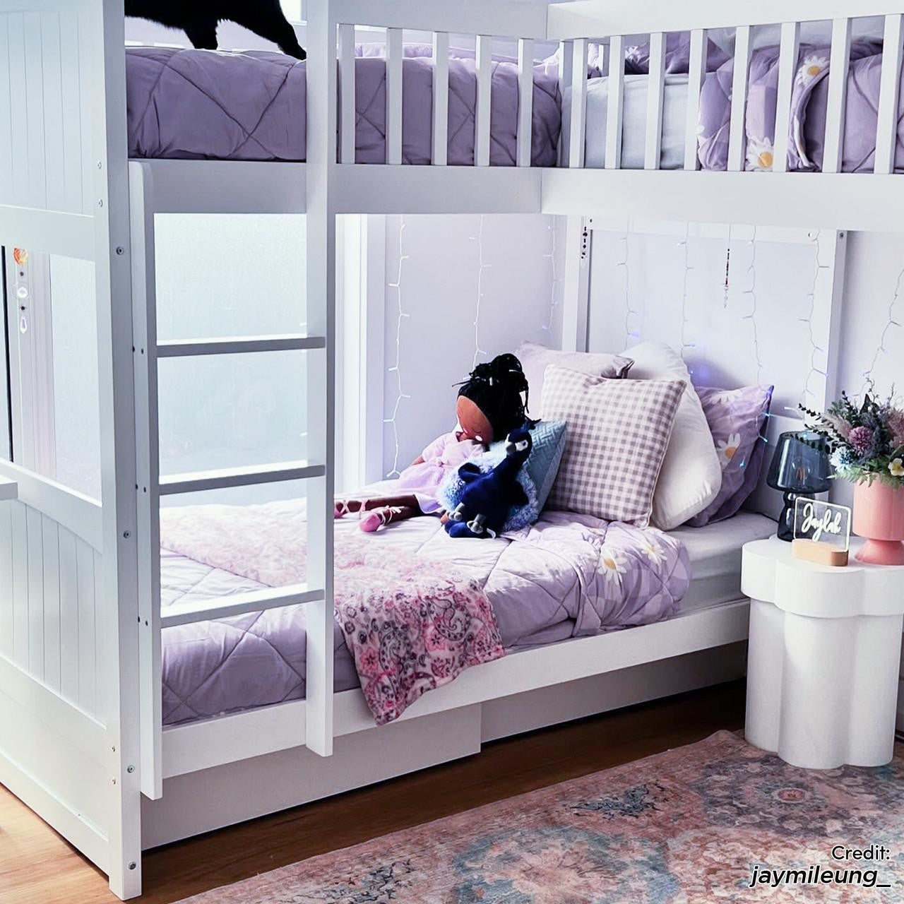 Lucy Solid Pine Wood Corner Triple Loft Bunk Bed in White, featuring a bunk with lavender bedding and a playful decor, surrounded by fairy lights, a side table with a lamp, and plush toys.