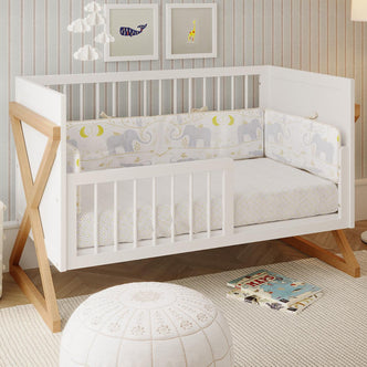 Melai Solid Pine Toddler Bed Rail Kit in white, installed on a crib with natural-wood “X” legs, surrounded by nursery decor including elephant-patterned bedding, a cloud mobile, and animal prints.