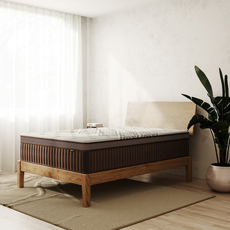Thalia Double 30cm Memory Foam Pocket Spring Mattress in Med positioned on a low wooden bed frame in a minimalist bedroom, complemented by a ribbed rug, sheer curtains, and a leafy plant.