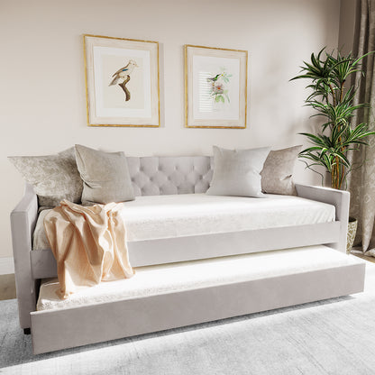 The Ovidia Single Velvet Daybed Sofa with Trundle in Light Grey features tufted upholstery, four neutral throw pillows, a peachy-beige throw, and framed botanical prints above, complemented by a leafy plant.
