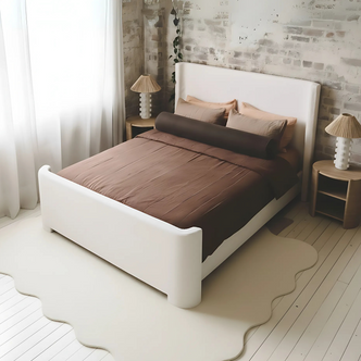 Palmira Queen Boucle Fabric Bed Frame in Cream White, featuring a low-profile upholstered design with a sleek sleigh silhouette, set against a white-washed brick wall and complemented by warm brown bedding.