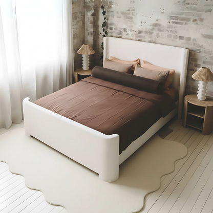 Palmira Queen Boucle Fabric Bed Frame in Cream White, featuring a low-profile upholstered design with a sleek sleigh silhouette, set against a white-washed brick wall and complemented by warm brown bedding.