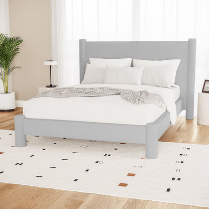 Rhoda Double Boucle Fabric Bed Frame in Light Grey with rounded posts, white bedding, knit throw, on a patterned rug. Flanked by a potted palm, side tables, and sheer curtains.