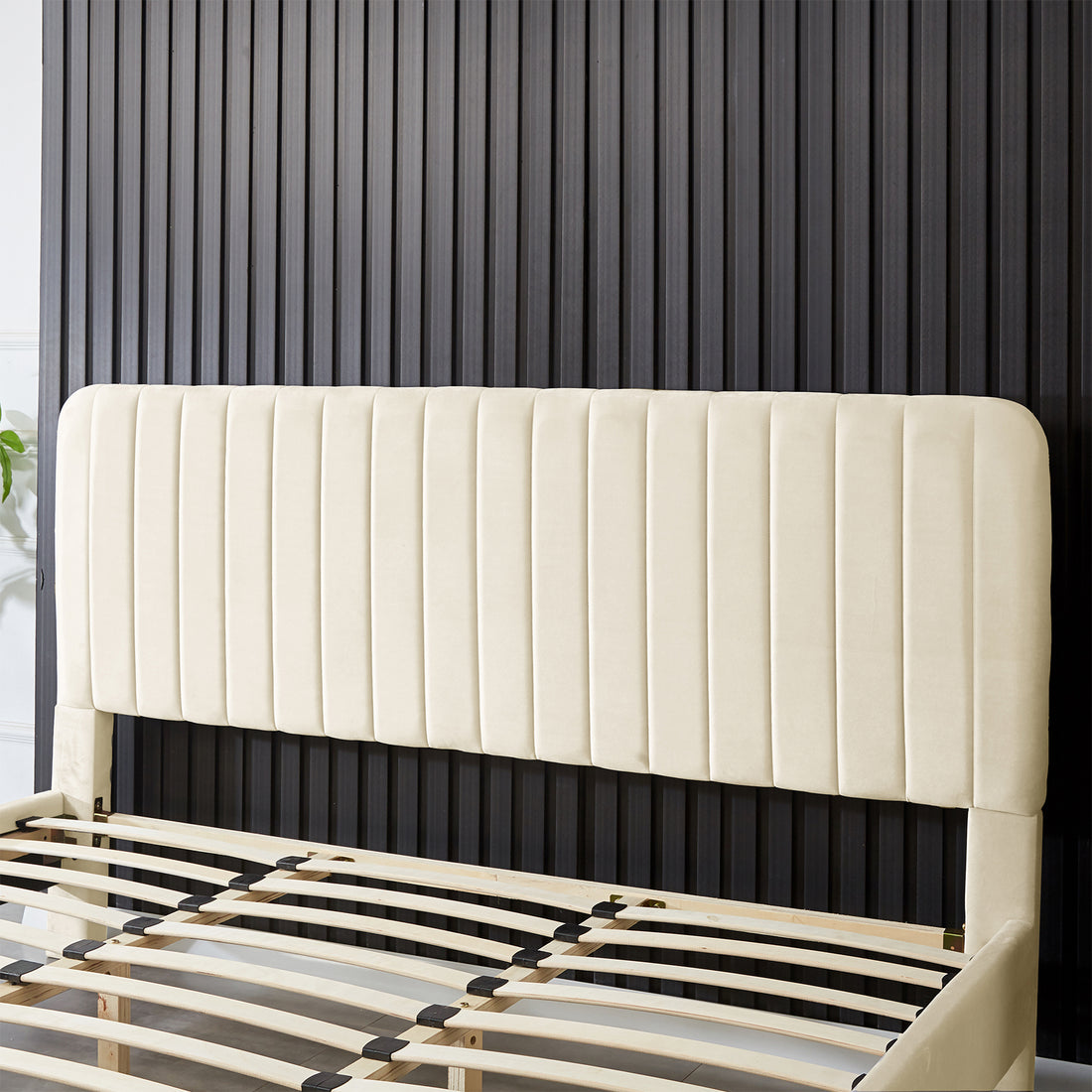 Seneca King Upholstered Velvet Bed – Sandy White features a tall, cream-colored headboard with vertical channeling and visible plywood slats, showcasing its elegant design against a dark ribbed wall.