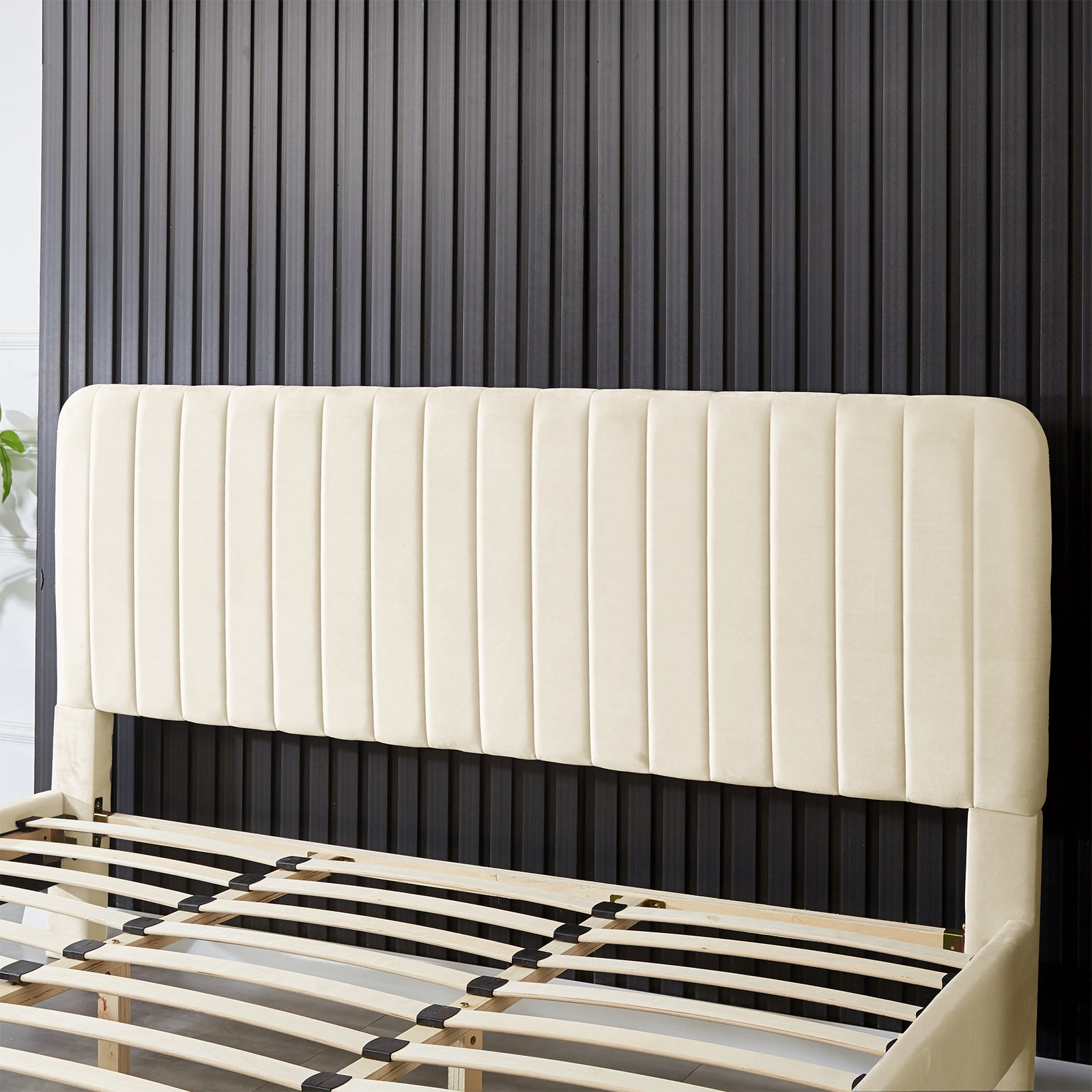 Seneca King Upholstered Velvet Bed – Sandy White features a tall, cream-colored headboard with vertical channeling and visible plywood slats, showcasing its elegant design against a dark ribbed wall.