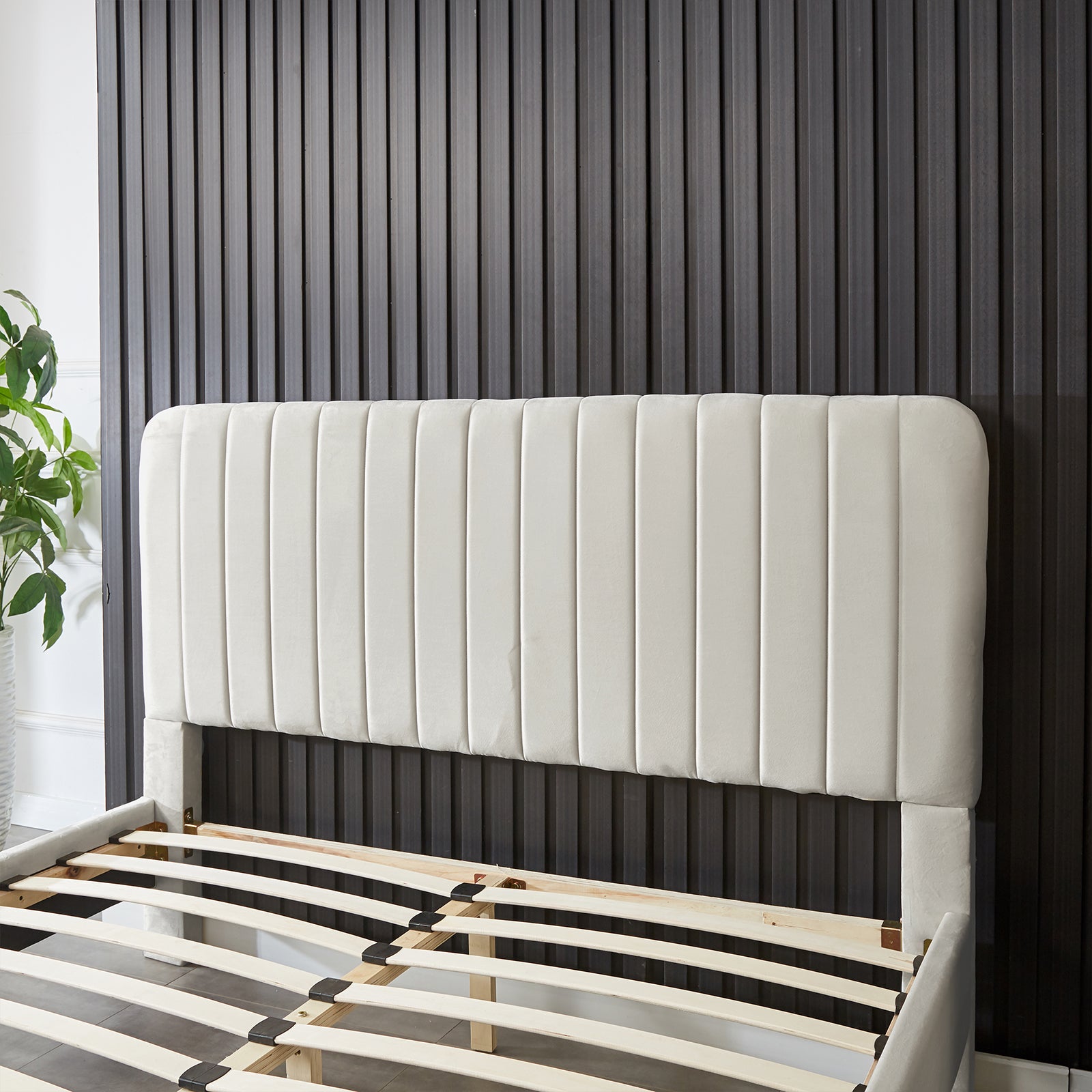 Seneca Queen Upholstered Velvet Bed – Slate features a tall padded headboard with subtle vertical stitching, wooden slats, and a potted houseplant beside it, set against a dark grooved wall.