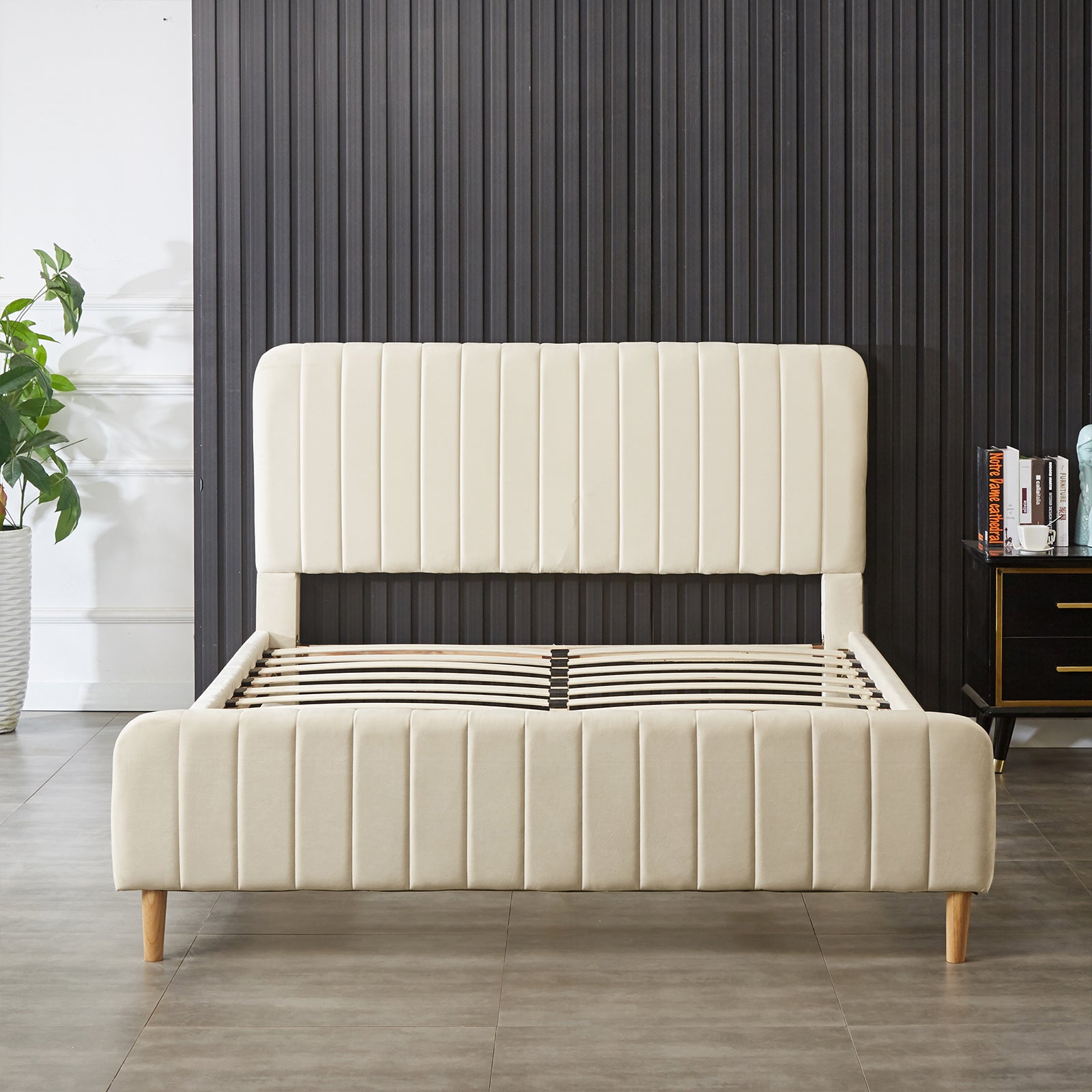 Seneca Queen Curved Velvet Fabric Bed Frame in Sandy White with vertically tufted headboard and footboard, wooden legs, and a support slat system, complemented by a potted plant and nightstand.