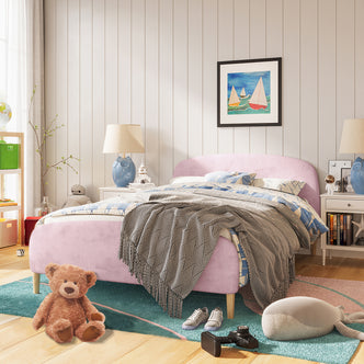 Theron Double Upholstered Velvet Bed in Light Pink, featuring a plush pink frame with a curved headboard, styled with blue-and-white bedding, gray throw, and surrounded by colorful and playful room accents.
