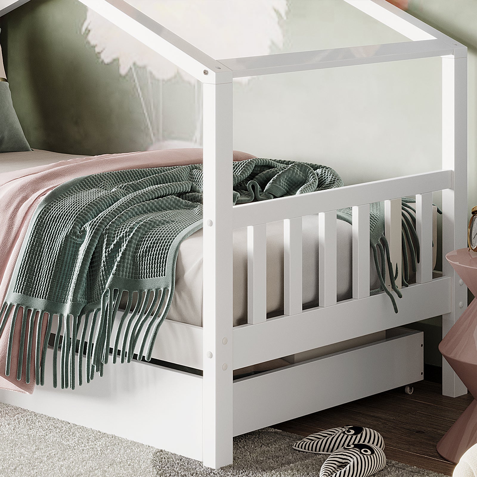 Tucker Solid Pine Timber Kids Bed with Drawers White, featuring a house-shaped frame, pink sheets, green throw, pull-out trundle, and nearby slippers on a rug over wood flooring.
