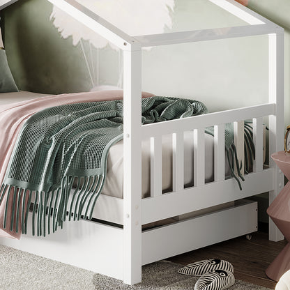 Tucker Solid Pine Timber Kids Bed with Drawers White, featuring a house-shaped frame, pink sheets, green throw, pull-out trundle, and nearby slippers on a rug over wood flooring.