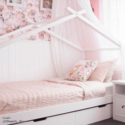 Tucker Solid Pine Timber Kids Bed with Drawers White features a house-shaped frame, soft pink bedding, tulle canopy, and two under-bed drawers in a pastel-pink children's bedroom setting.