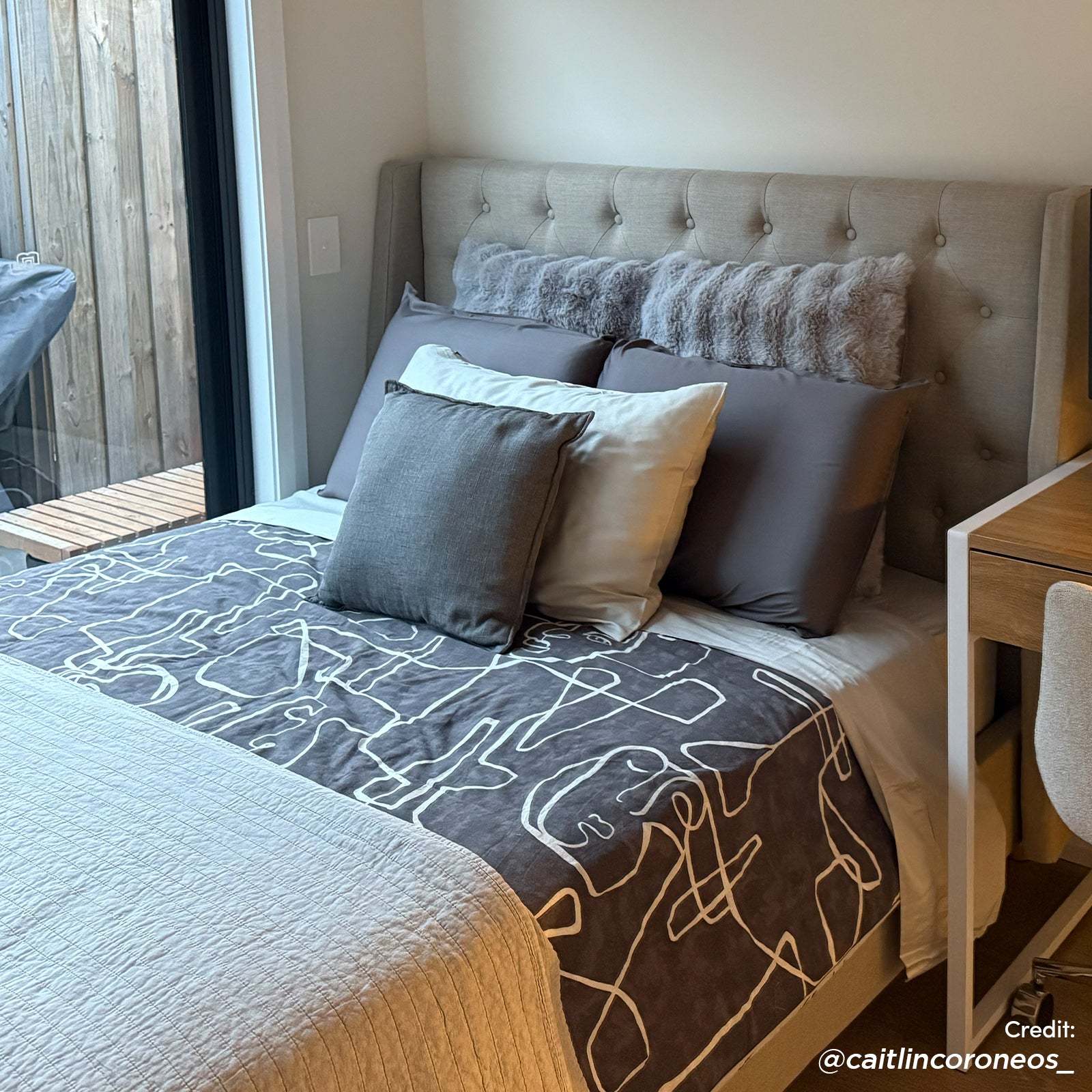 Vilania Double Linen Fabric Bed Frame with Buttons in Grey, featuring a light-grey, button-tufted headboard and charcoal-grey duvet, complements the modern bedroom setup with a desk and glass door.