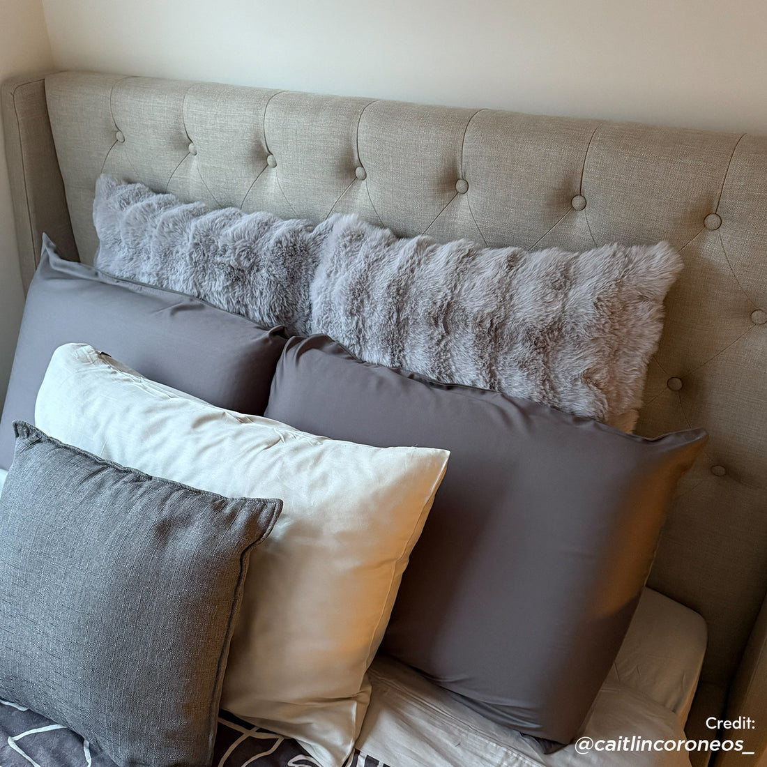 Vilania Double Linen Fabric Bed Frame with Buttons in Grey features a light-beige, button-tufted headboard. Highlighted by a layered arrangement of gray and ivory pillows, presenting a cozy, neutral bedroom aesthetic.