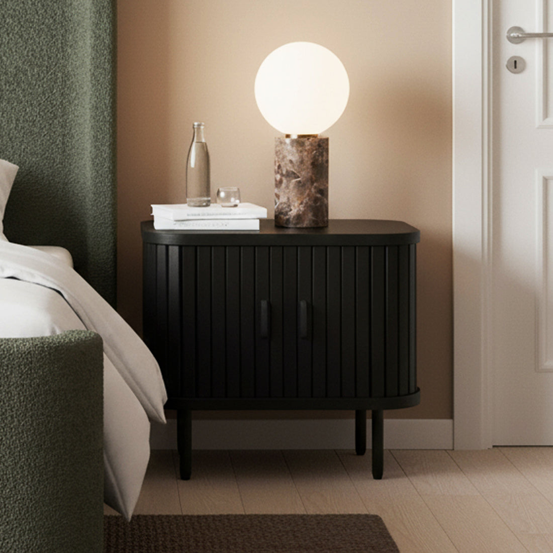 Aaron 50cm Fluted Curved Bedside Table in Black with sliding doors, displaying a glass water carafe, white books, and a marble lamp on top, set in a modern bedroom corner.