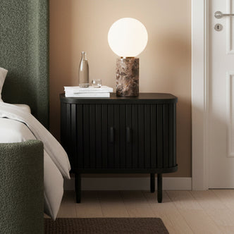 Aaron 50cm Fluted Curved Bedside Table in Black with sliding doors, displaying a glass water carafe, white books, and a marble lamp on top, set in a modern bedroom corner.