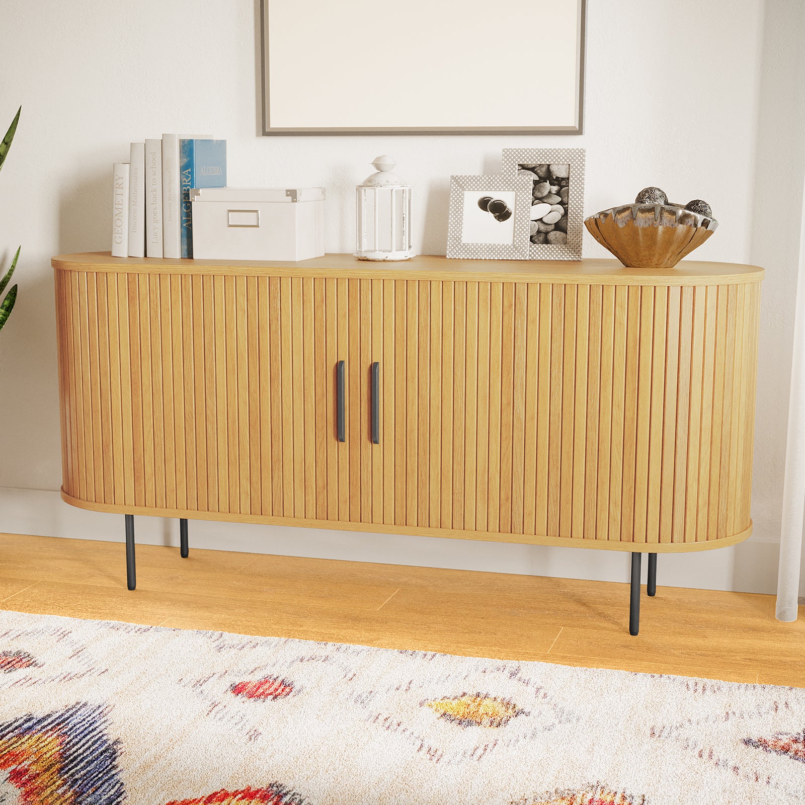 The Aaron 140cm Curved Sideboard Buffet - Natural features a mid-century design with rounded front, fluted wooden doors, slim black metal legs, and is topped with books, decor, and framed photos.