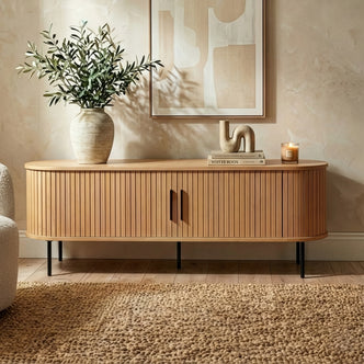 Aaron 160cm Fluted Curved Entertainment TV Unit in Natural with vertical slats, slim black metal legs, topped with a vase, books, abstract sculpture, and a candle, enhancing a warm, minimal decor.