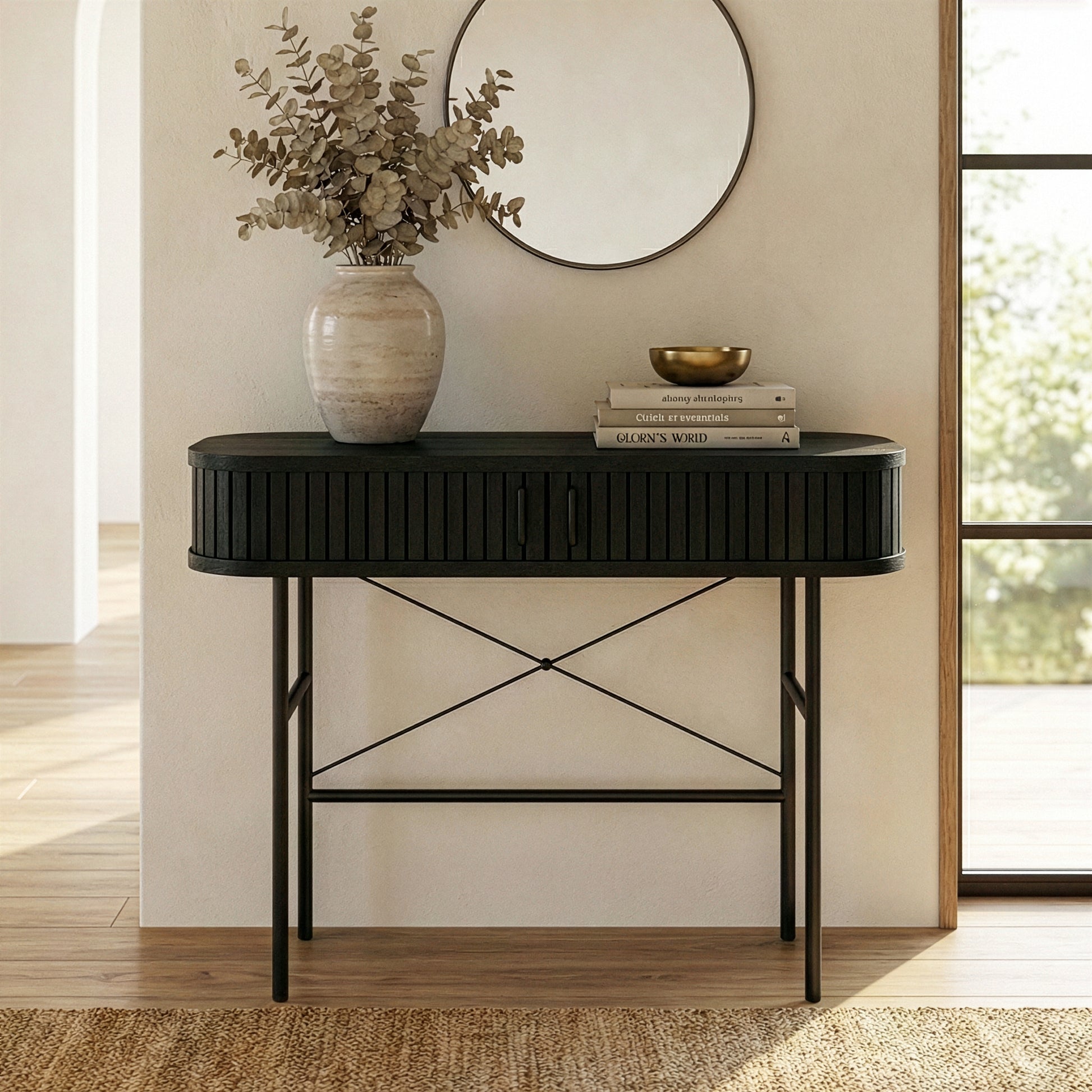 Aaron 100cm Fluted Curved Hallway Table in Black with sliding doors, adorned with a rustic vase, eucalyptus, books, and a brass-tone bowl, below a round mirror in a softly lit room.
