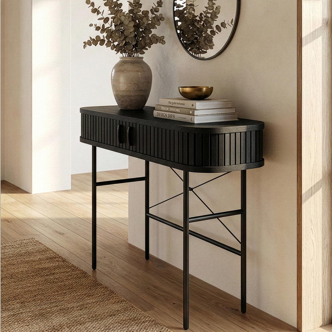 Aaron 100cm Fluted Curved Hallway Table in Black with slender metal legs features a stylish mirror above, a ceramic vase with eucalyptus, books, and a brass bowl, creating a chic entryway vignette.
