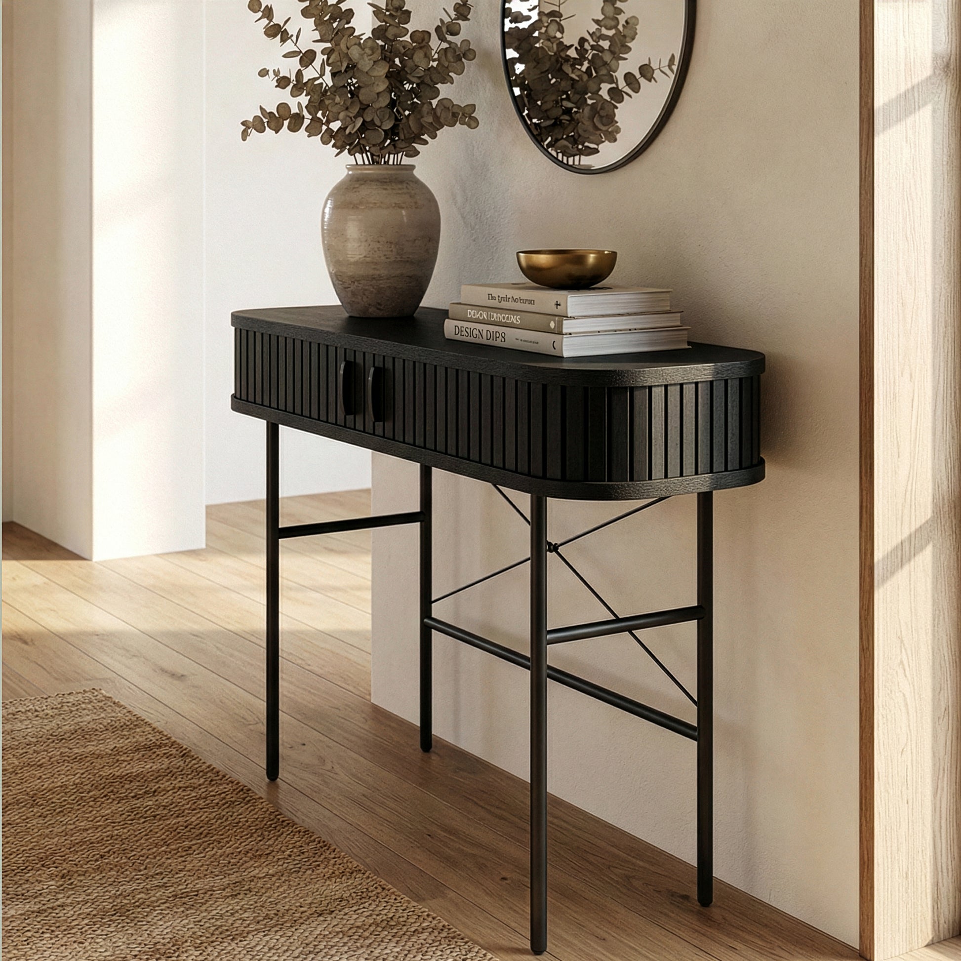 Aaron 100cm Fluted Curved Hallway Table in Black with slender metal legs features a stylish mirror above, a ceramic vase with eucalyptus, books, and a brass bowl, creating a chic entryway vignette.