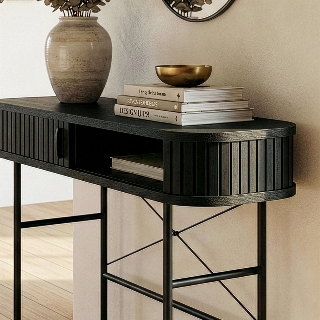 Aaron 100cm Fluted Curved Hallway Table in Black with a sleek wooden top, slender metal legs, adorned with design books, a brass bowl, and a vase with dried foliage, under a round mirror.
