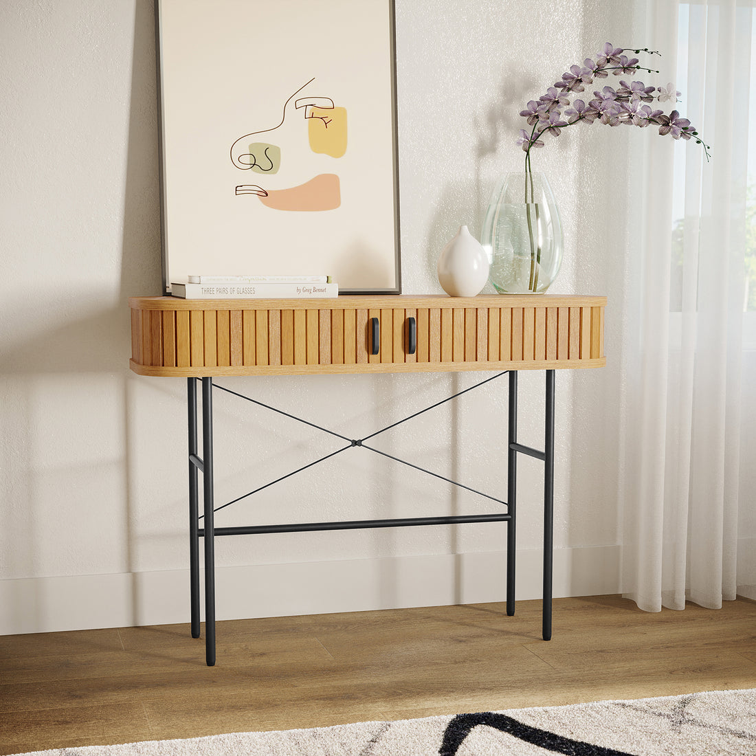 Aaron 100cm Hallway Table with Sliding Doors - Natural, featuring a slatted wood top, black metal legs, two sliding doors, with books, a framed print, and vases with purple blossoms on top.