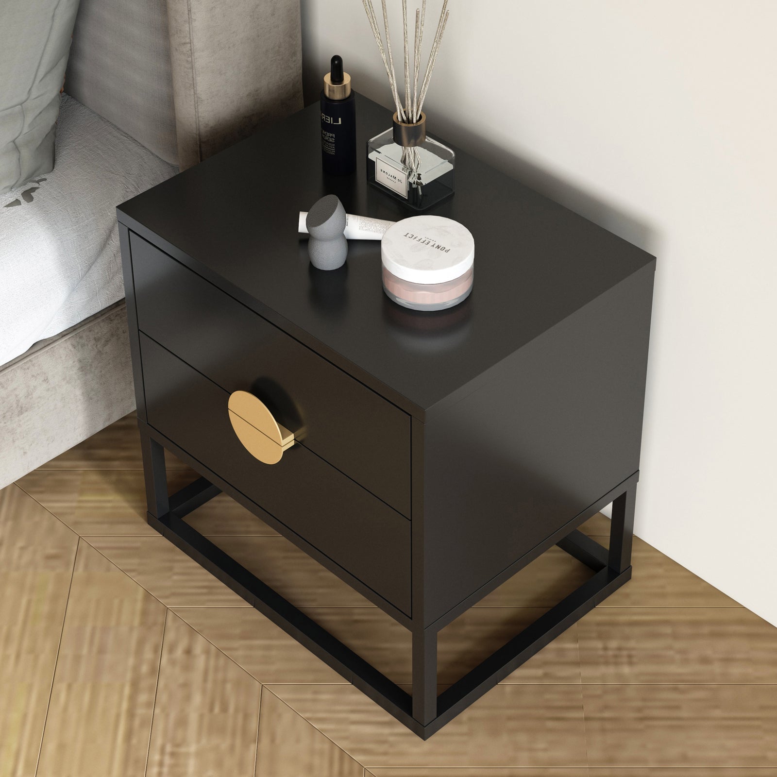 Abigail 55cm 2-Drawer Bedside Table - Black/Gold, features a sleek metal frame, gold handles, on a parquet floor, topped with skincare items and a makeup sponge, next to a bed.