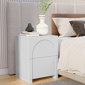 Archen 2 Drawer Arch Detail Bedside Table in White with books, vases, and a green twig on top, beside a bed with neutral linens, against a vertical-slatted wall.