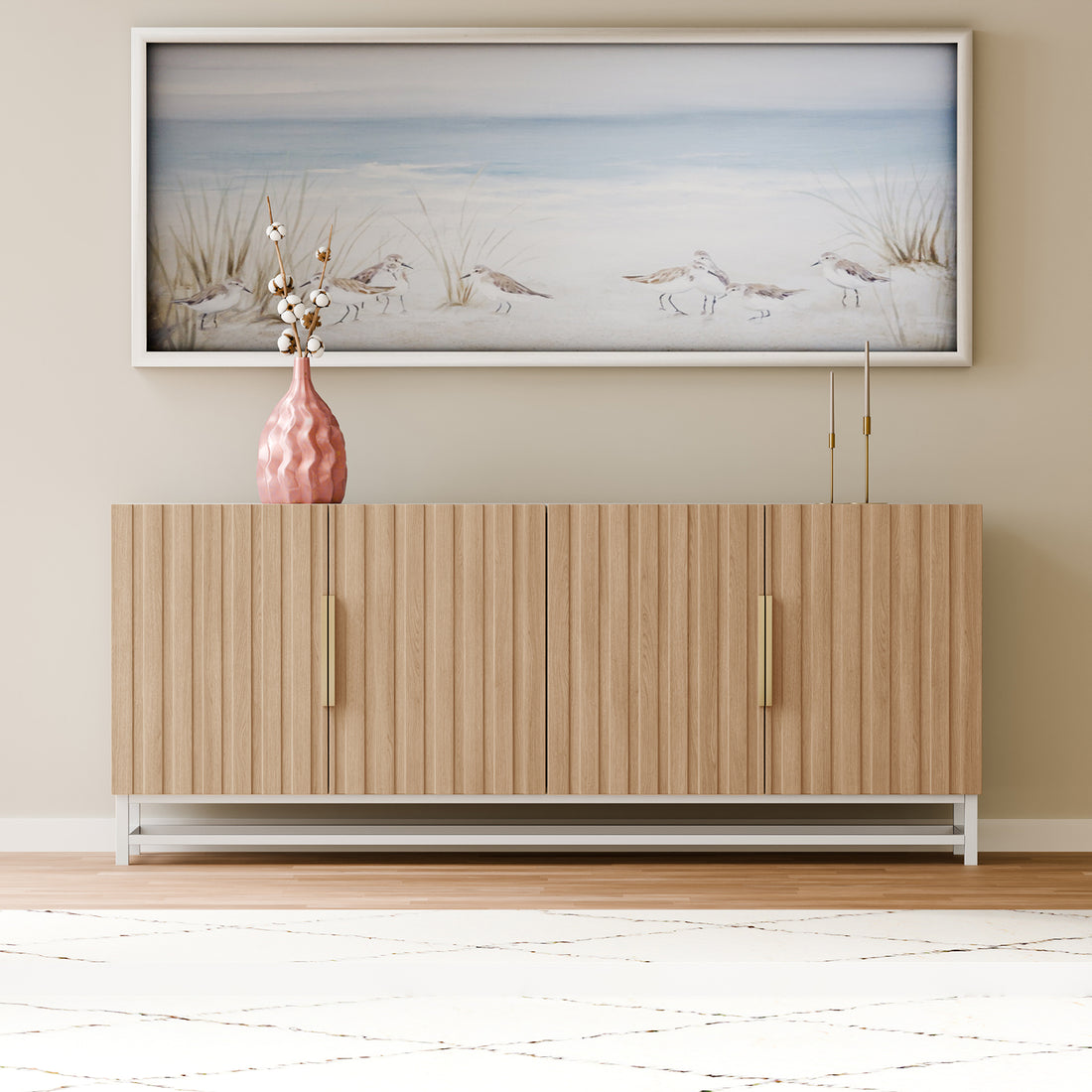 Argus 180cm Fluted Storage Buffet Sideboard in Natural features a light-wood design with vertical slats and slim brass handles, topped with a pink vase, gold candleholders, and a beach scene print above.