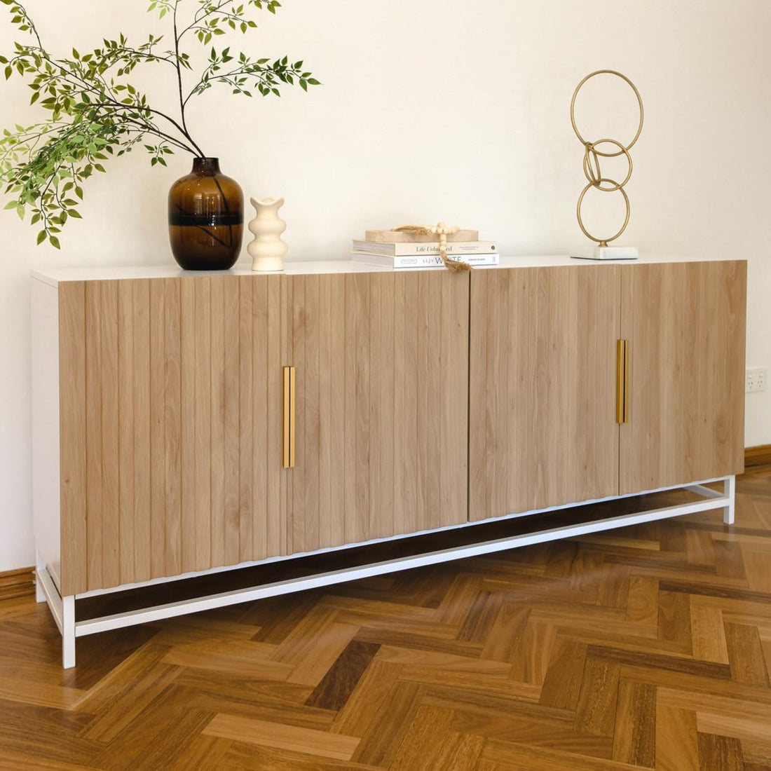 Argus 180cm Fluted Storage Buffet Sideboard in Natural with white metal base, four vertical-slatted wooden doors with brass handles, topped with amber vase, cream ceramic vase, books, and gold sculpture.