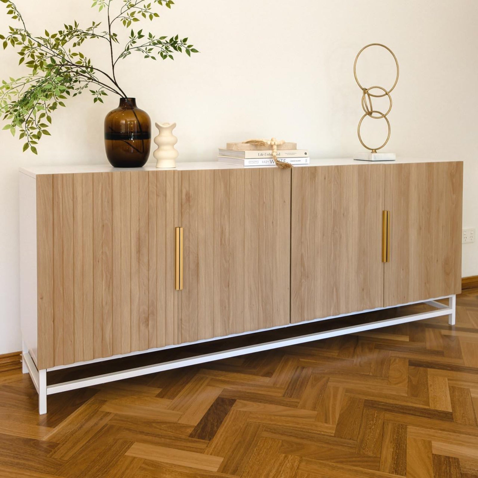 Argus 180cm Fluted Storage Buffet Sideboard in Natural with white metal base, four vertical-slatted wooden doors with brass handles, topped with amber vase, cream ceramic vase, books, and gold sculpture.