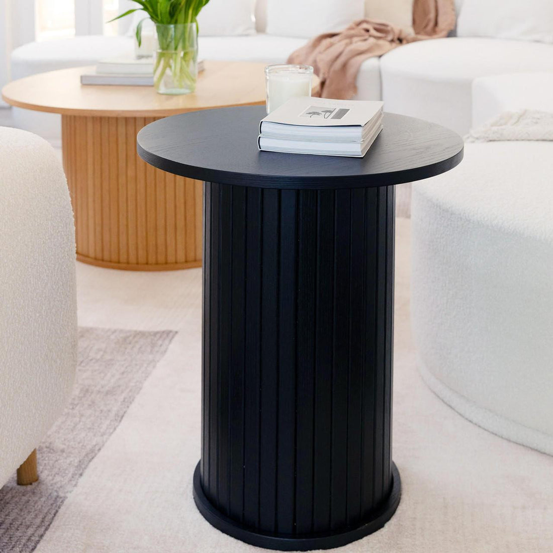 The Booker 50cm Round Fluted Oak Side Table in black features a sleek cylindrical design with vertical fluting, topped with books and a candle, enhancing a modern living room's elegance.