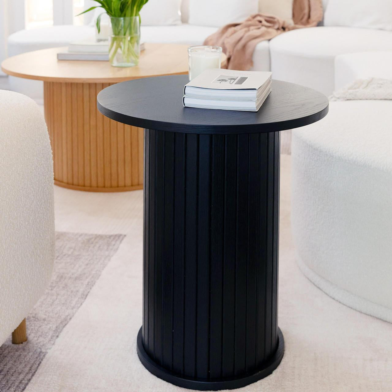The Booker 50cm Round Fluted Oak Side Table in black features a sleek cylindrical design with vertical fluting, topped with books and a candle, enhancing a modern living room's elegance.