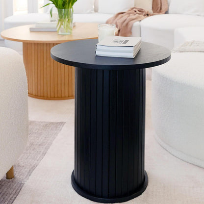The Booker 50cm Round Fluted Oak Side Table in black features a sleek cylindrical design with vertical fluting, topped with books and a candle, enhancing a modern living room's elegance.