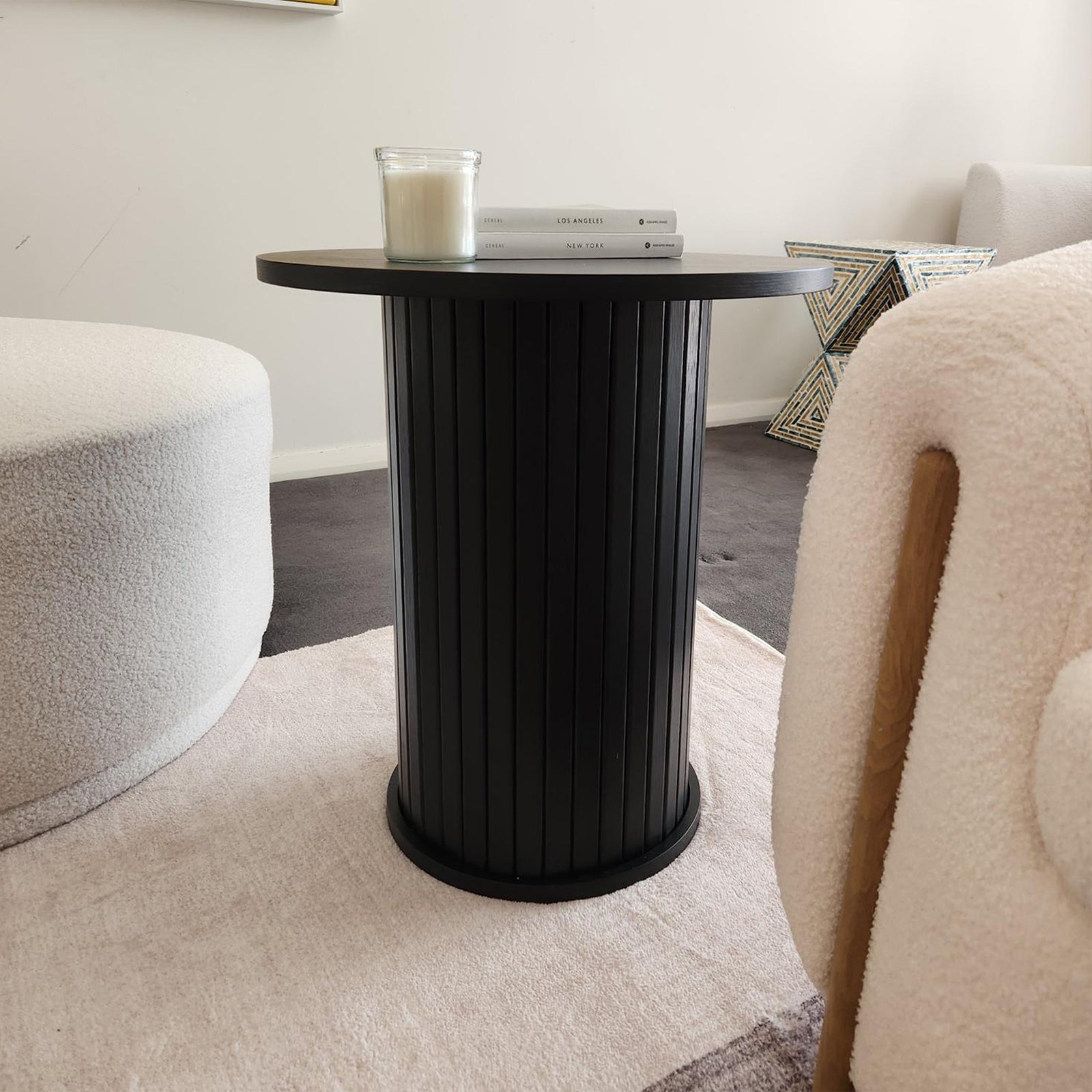 Booker 50cm Round Fluted Oak Side Table in Black, featuring a ribbed design, holds a glass-jar candle and books. Positioned between a boucle ottoman and armchair on a neutral rug.