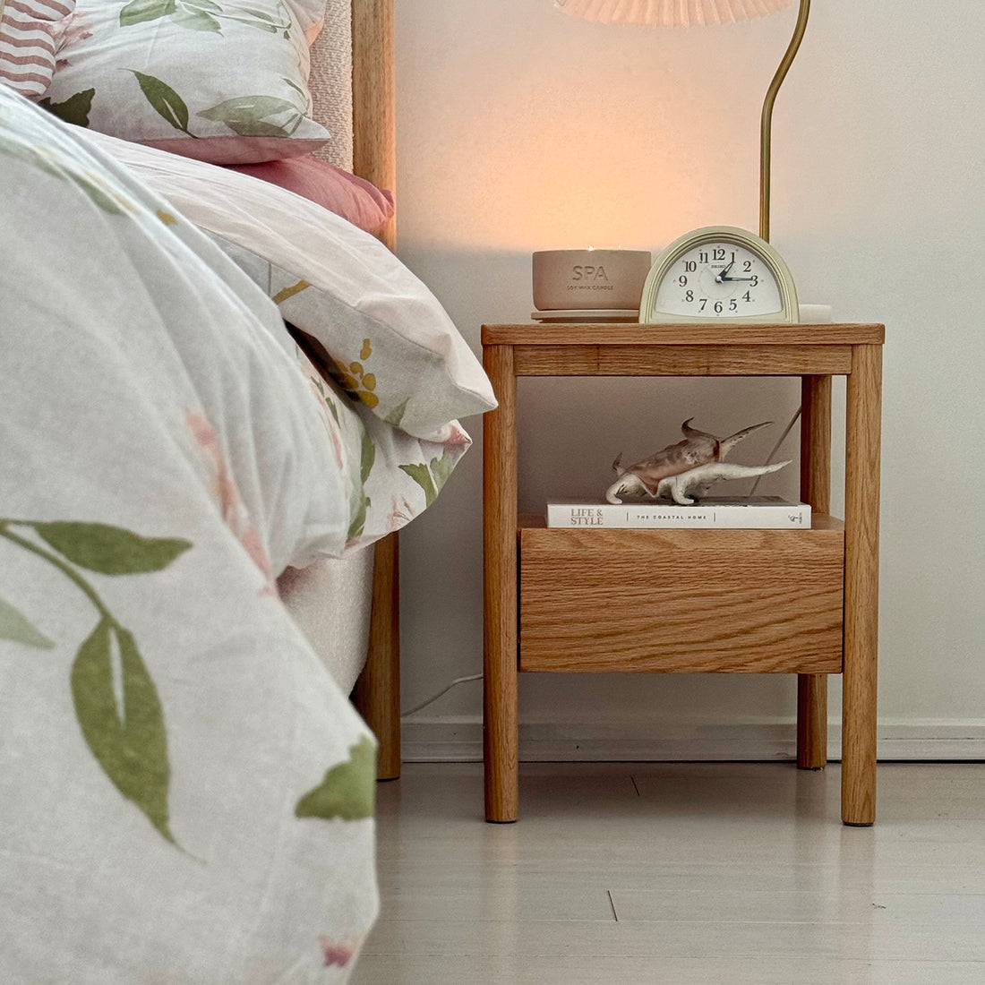 Brett Solid Oak Bedside Table - Natural next to a bed, featuring a drawer and open shelf, crafted from solid oak with a natural finish, enhanced by a lamp on the nightstand.