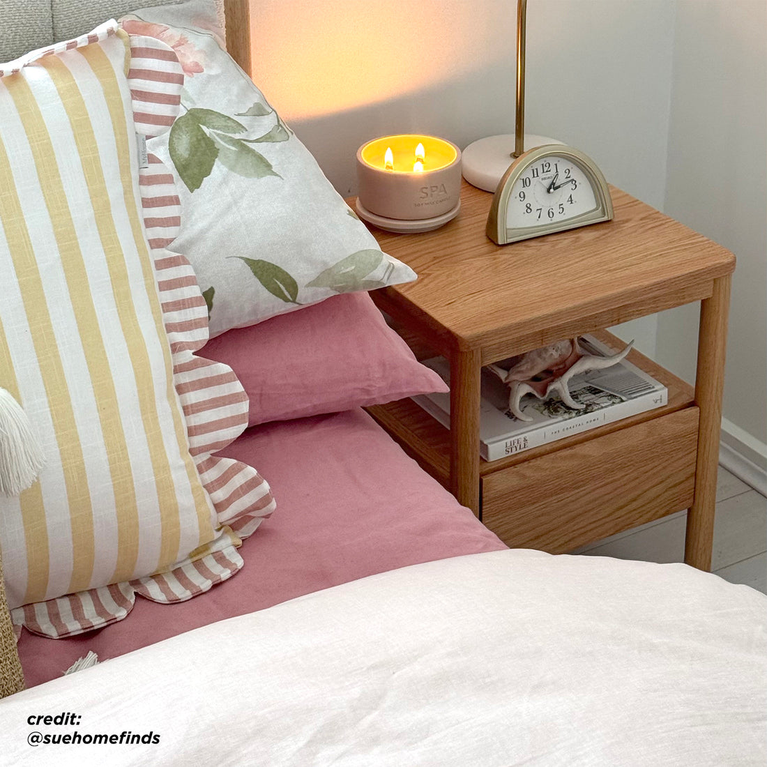 Brett Solid Oak Bedside Table - Natural displayed with a smooth, natural wood finish, featuring a spacious drawer and open shelf, set in a cozy indoor environment with plush pillows and a clock.