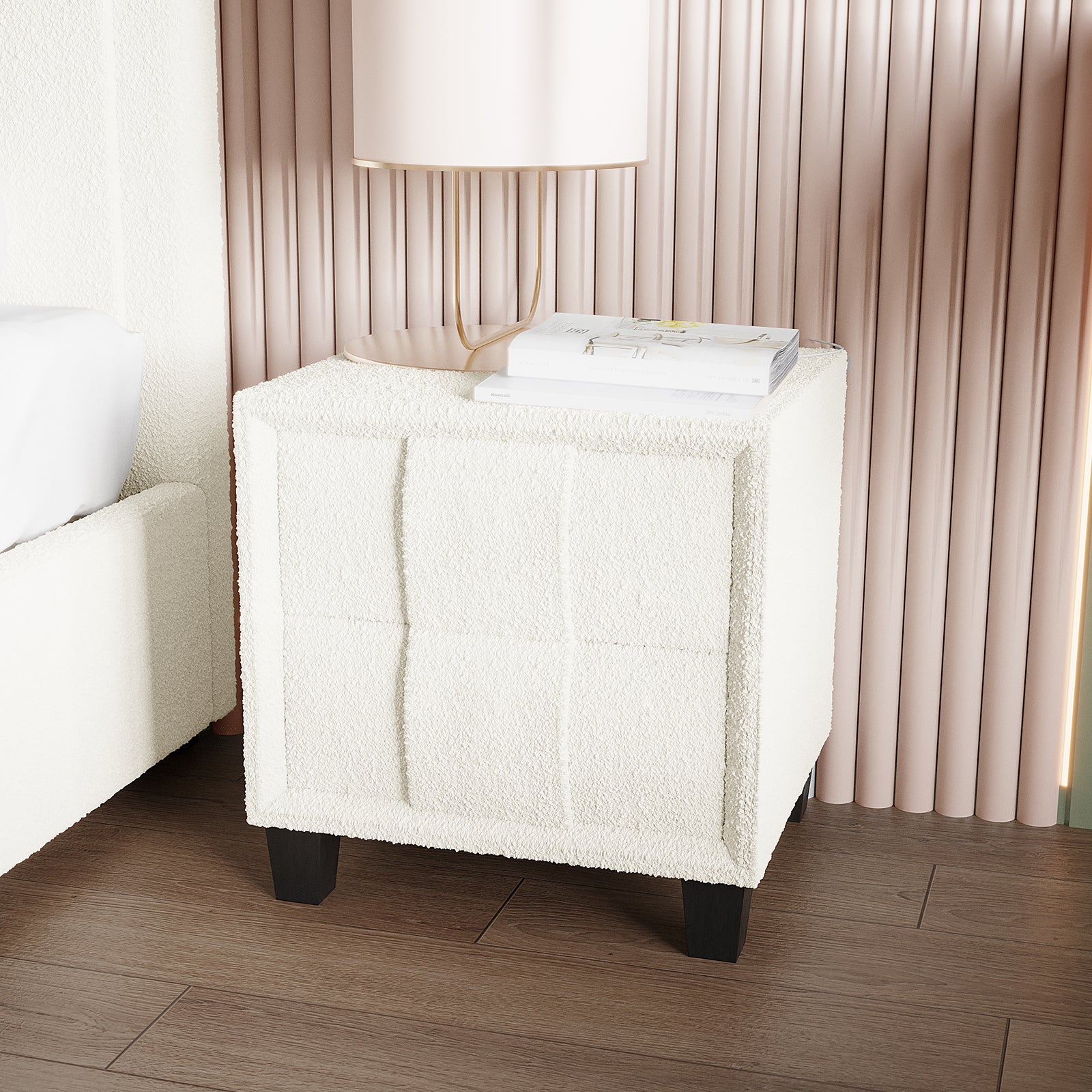 Crossandra Boucle Bedside Table in Cream White, upholstered in soft boucle fabric with dark wooden legs, topped with a pale lamp and white books, against blush-pink slatted wall.