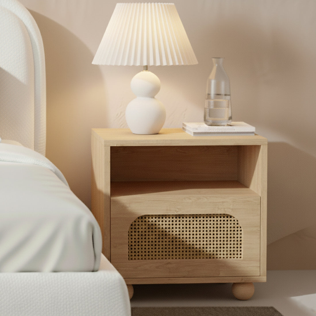 Dialga 45cm Rattan Bedside Table with Storage in Natural, featuring a woven-rattan-front drawer, open shelf, and light-wood finish, complemented by a white lamp and books on top.