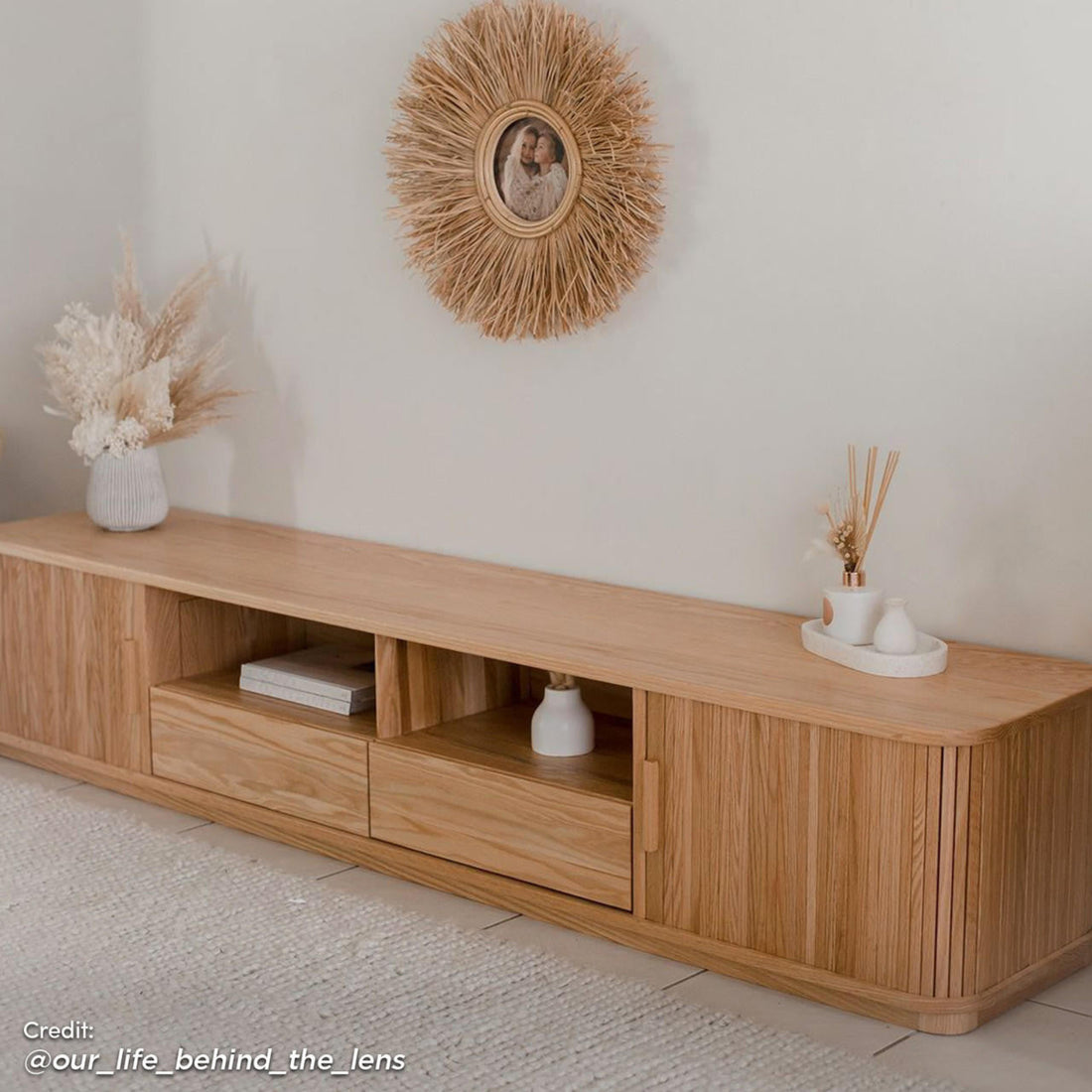 Eddie 200cm Hardwood Oak TV Entertainment Unit in Natural, featuring a minimalist design with sliding doors, drawers, and open cubbies, adorned with pampas grass and a sunburst rattan-framed photograph above.