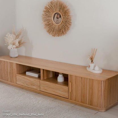 Eddie 200cm Hardwood Oak TV Entertainment Unit in Natural, featuring a minimalist design with sliding doors, drawers, and open cubbies, adorned with pampas grass and a sunburst rattan-framed photograph above.