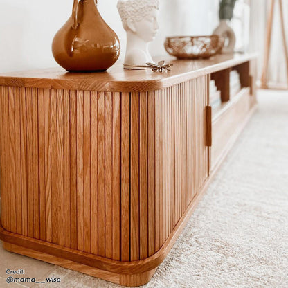 Eddie 200cm Hardwood Oak TV Entertainment Unit in Natural with slatted doors on pale carpet, topped with a ceramic vase, sculptural bust, metal trinket, and woven basket, showcasing minimalist design.