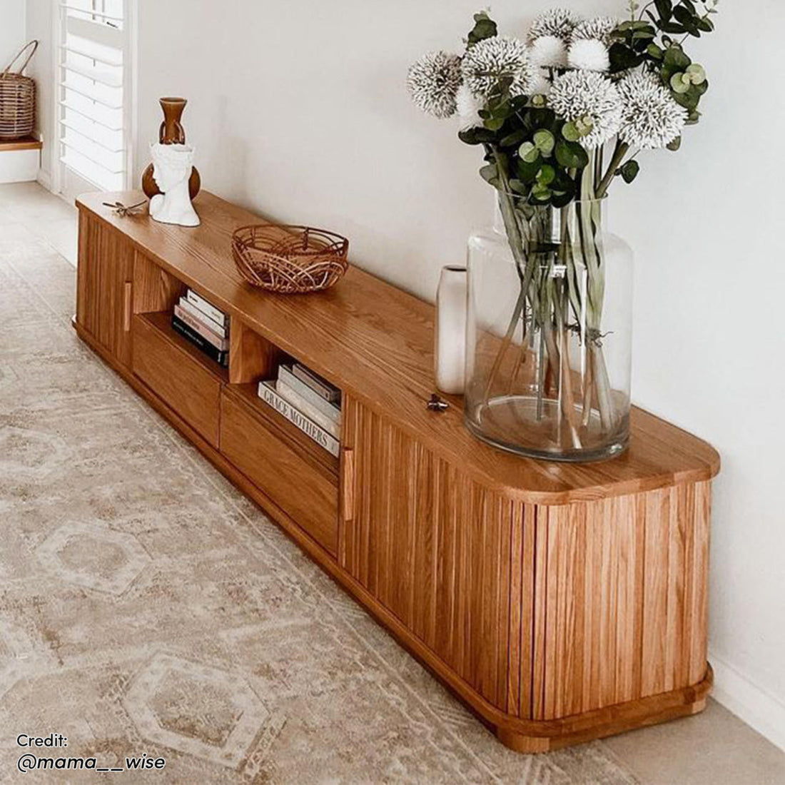 Eddie 200cm Hardwood Oak TV Entertainment Unit in Natural, featuring open cubbies with books, topped with decorative vases, a bust, and a woven basket, against a pale wall and geometric rug.