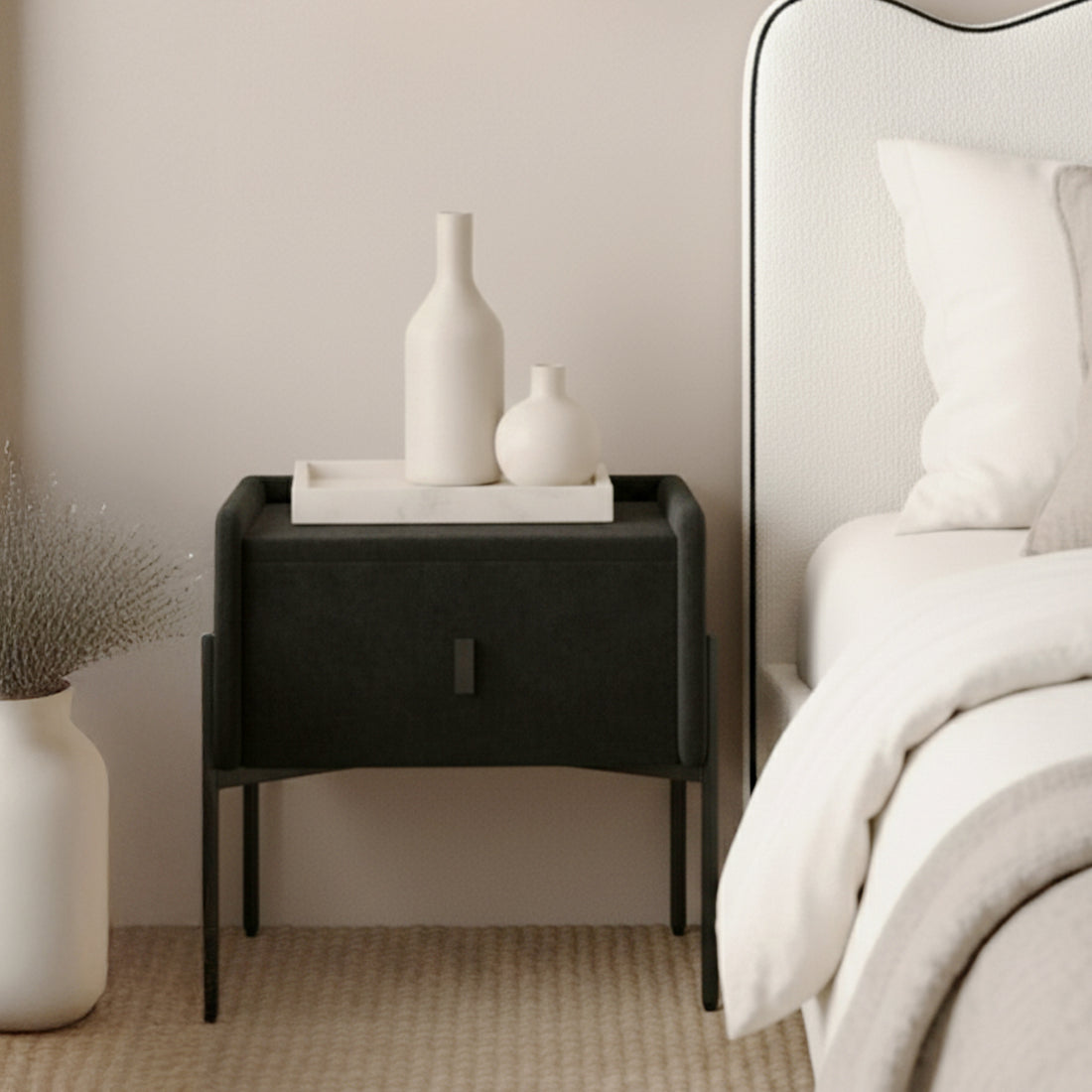 Eliz Velvet Fabric Bedside Table in Black with slender legs, topped with a white tray and ceramic vases, beside a bed with white bedding, exuding a minimalist, contemporary aesthetic.